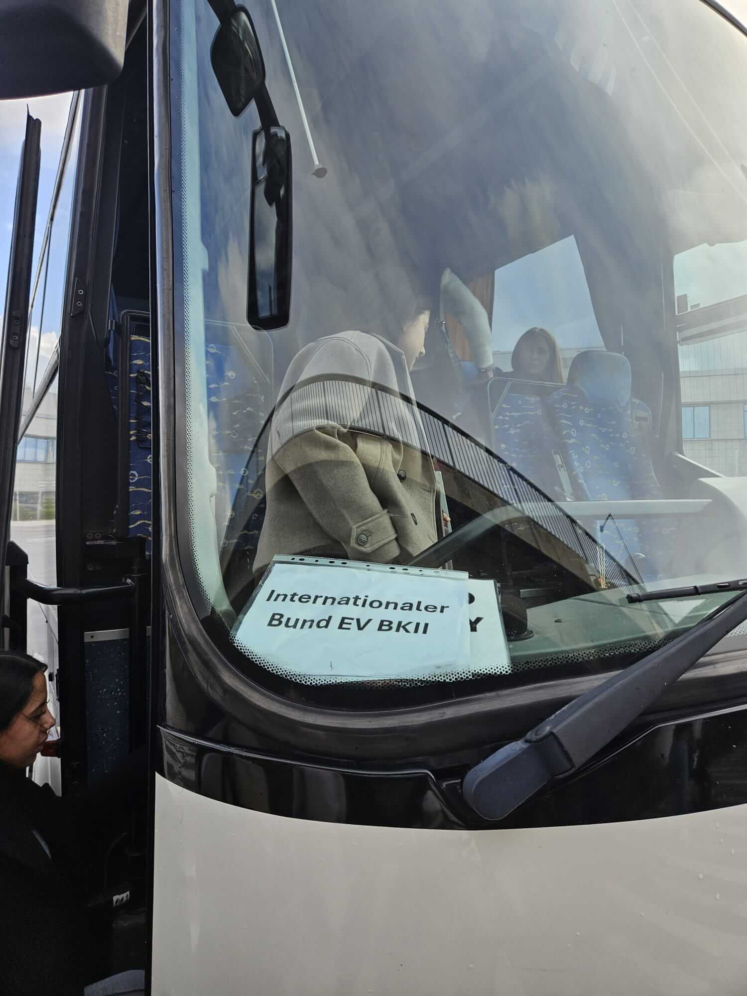 Ein Bus mit dem Schild "Internationaler Bund EV BKII" wird bestiegen, während Passagiere durch die Frontscheibe sichtbar sind.