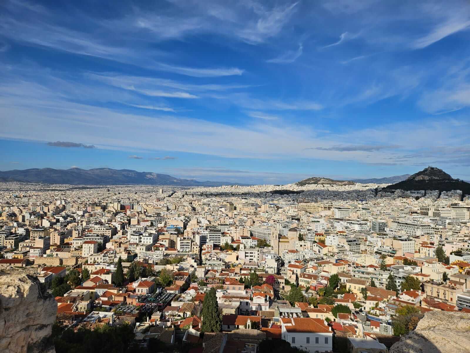Panoramablick über Athen unter blauem Himmel, mit dicht bebauten Häusern und Hügeln im Hintergrund.