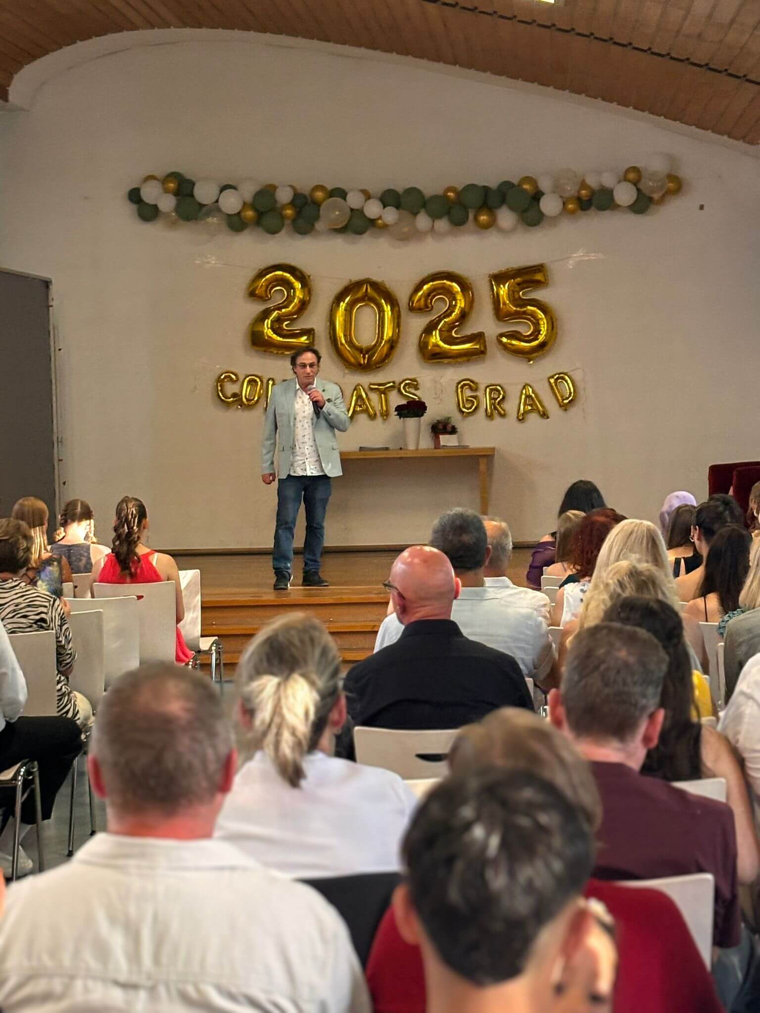 Ein Redner steht auf einer Bühne vor einer "2025 Congrats Grad"-Dekoration, während das Publikum aufmerksam zuhört.
