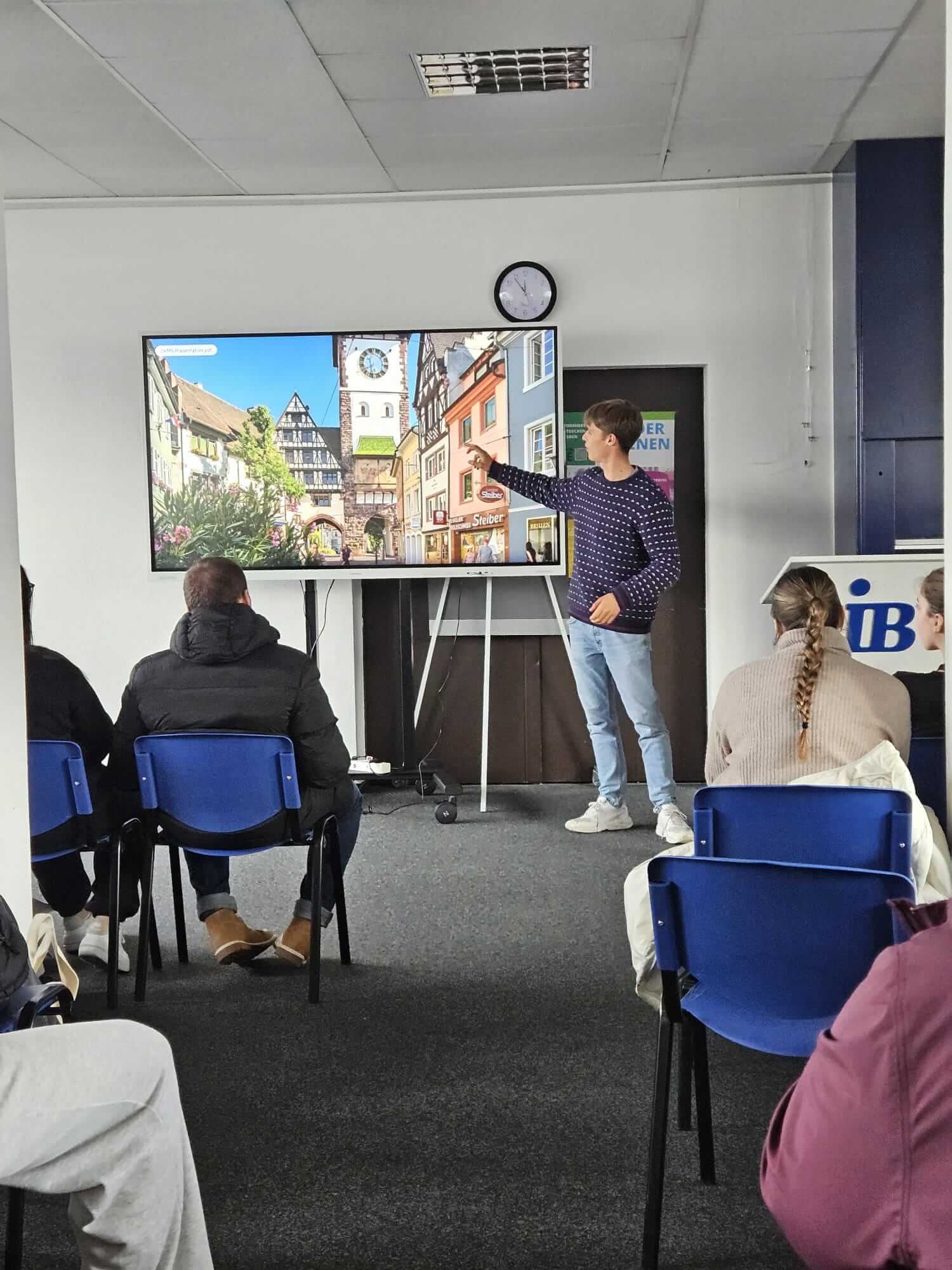 Ein junger Mann hält eine Präsentation vor einer Gruppe, während er auf ein Bild einer malerischen Stadt auf dem Bildschirm zeigt.