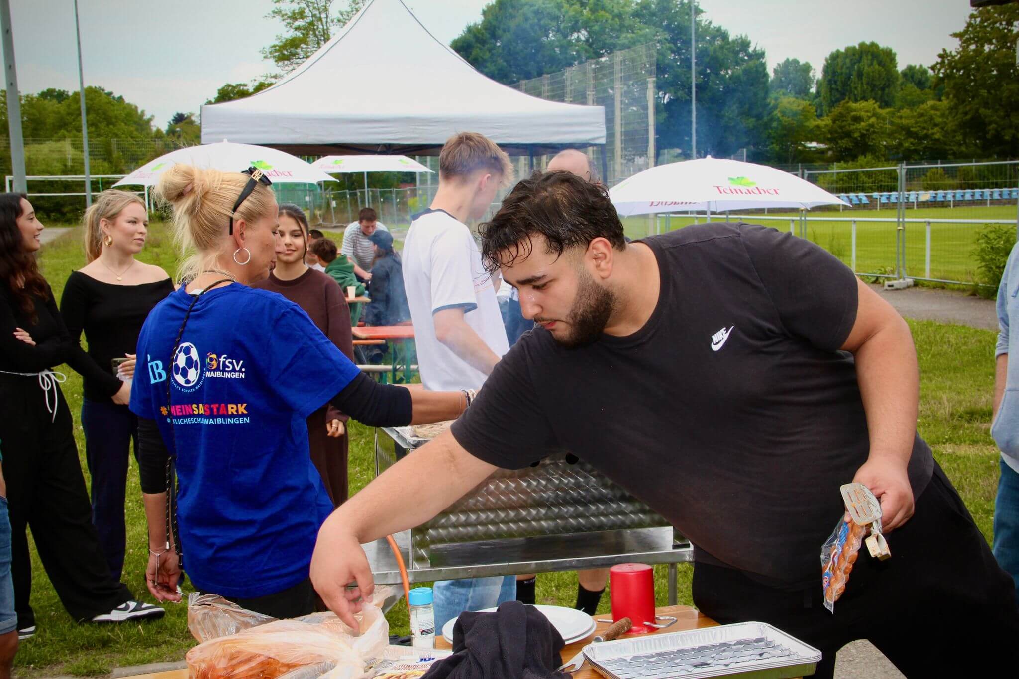 Ein Mann bereitet Essen auf einem Grill bei einem sommerlichen Fußballfest im Freien zu, umgeben von geselligen Menschen.