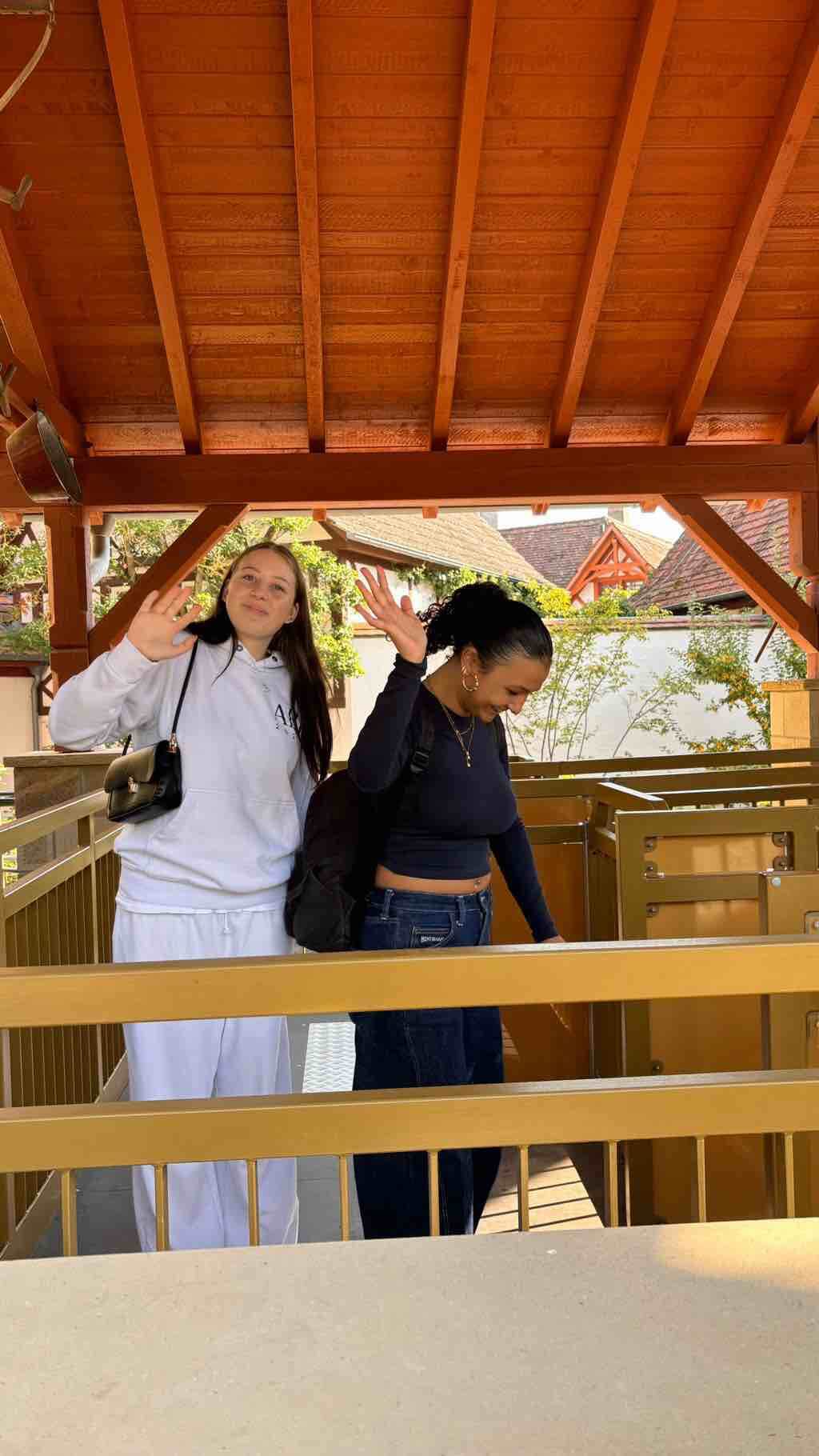 Zwei Frauen winken fröhlich unter einem hölzernen Pavillon in einer sonnigen, charmanten Umgebung.