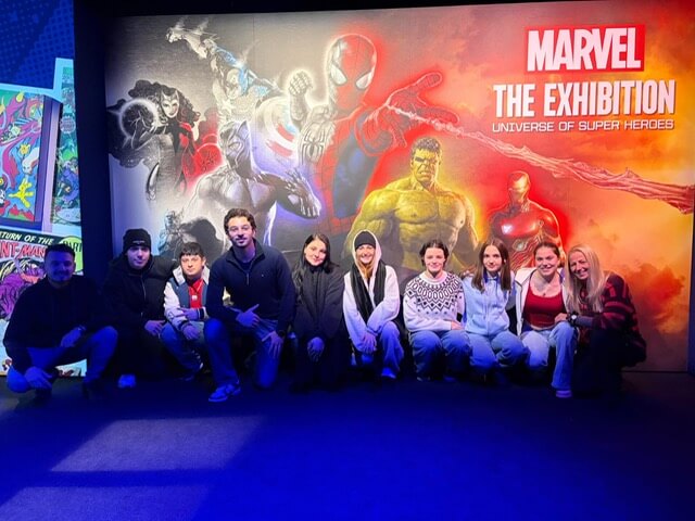 Eine Gruppe von Menschen posiert vor einem Marvel-Ausstellungsplakat mit Superheldenmotiven.